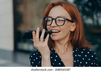 Close up of young smiling red-haired business woman holds mobile phone and using virtual digital voice assistant while standing outdoors, happy female talks on speakerphone on smartphone - Powered by Shutterstock