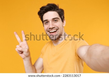 Close up young smiling unshaved friendly caucasian man 20s wear basic blank print design t-shirt doing selfie shot on mobile phone show victory sign isolated on yellow color background studio portrait