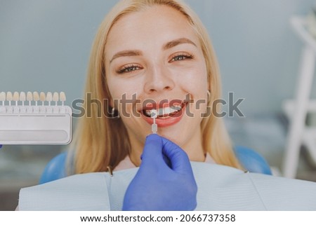 Close up young smiling woman choose veneers enamel color palette with stomatologist compare teeth shade sit at dentist office chair indoor light modern cabinet Healthcare enamel whitening treatment.