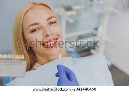 Close up young smiling woman choosing veneers enamel color palette with stomatologist compare teeth shade sit at dentist office chair indoor light modern cabinet Healthcare enamel whitening treatment
