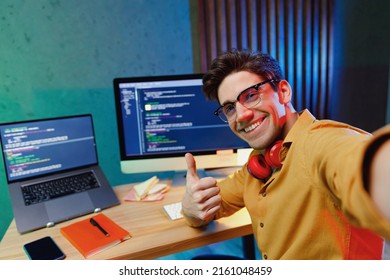 Close up young software site reliability engineer IT programmer man in shirt work at home write code on laptop pc computer do selfie shot pov on mobile phone show thumb up Program development concept