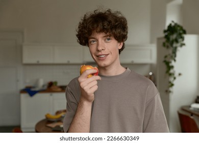 Close up of young thoughtful man with curly brown hair wearing casual t-shirt smiling at the camera looking away and eating an apple holding it in hands enjoying fruit in light kitchen at home.   - Powered by Shutterstock