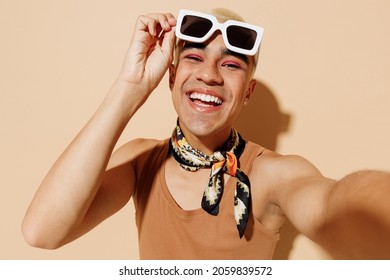 Close up young trendy fashionable blond latin gay man in beige tank shirt neck scarf take off sunglasses do selfie shot pov on mobile phone look aside isolated on plain light ocher background studio - Powered by Shutterstock