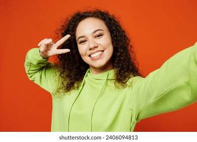 Close up young woman of African American ethnicity wear green hoody casual clothes do selfie shot pov mobile cell phone cover eye with v-sign isolated on plain red orange background. Lifestyle concept