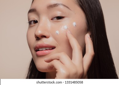 Close up of a young woman applying moisturizer to her face. Asian woman looking happy while following skincare regime.