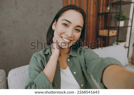 Close up young woman of Asian ethnicity wears casual clothes doing selfie shot pov on mobile cell phone sits on grey sofa couch stay at home hotel flat rest relax spend free spare time in living room