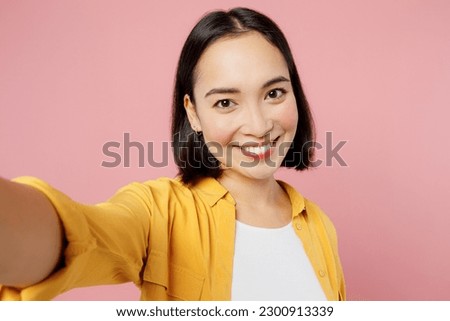Close up young woman of Asian ethnicity wears yellow shirt white t-shirt doing selfie shot pov on mobile cell phone isolated on plain pastel light pink background studio portrait. Lifestyle concept