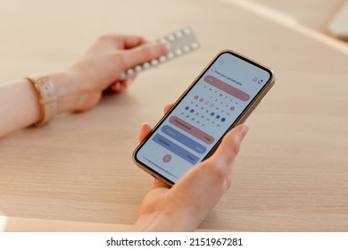 Close up of young woman calculating menstrual cycle using mobile app and holding birth control pills