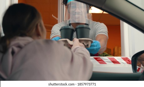 Close Up Of Young Woman In Car Receiving Coffee In Drive Thru Fast Food Restaurant. Cafe Staff In Protective Face Shield And Gloves Serving Takeaway Order For Driver In Delivery Window