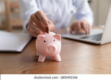 Close up young woman doctor putting coin into pink piggy bank, sitting at work desk, using laptop, hospital budget and accounting, finances, medical insurance, healthcare money savings concept