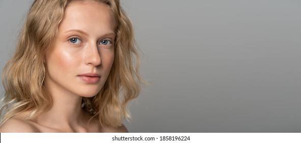 Close up of young woman face with blue eyes, curly natural blonde hair and eyebrows, has no makeup, looking at camera. Girl with perfect fresh clean skin over studio grey background with copy space.