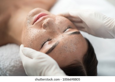 Close up of young woman getting facial skin treatment at spa. Cosmetician hands applying peeling mask on her face