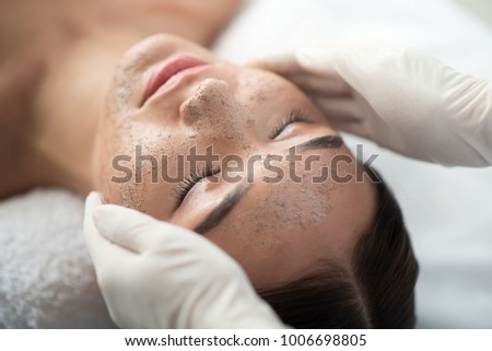 Close up of young woman getting facial skin treatment at spa. Cosmetician hands applying peeling mask on her face