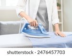 Close up young woman hand use electric steam, hot iron press pile stripe shirt clothes on ironing board, housework after hygiene laundry at home. Housekeeping lifestyle, household of chores concept.