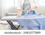 Close up young woman hand use electric steam, hot iron press pile stripe shirt clothes on ironing board, housework after hygiene laundry at home. Housekeeping lifestyle, household of chores concept.