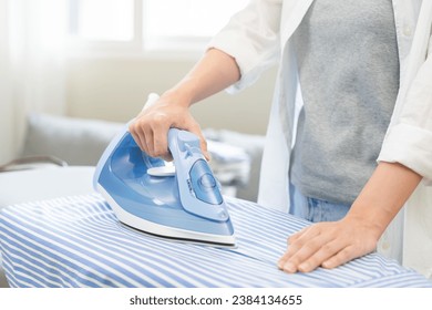 Close up young woman hand use electric steam, hot iron press pile stripe shirt clothes on ironing board, housework after hygiene laundry at home. Housekeeping lifestyle, household of chores concept.
