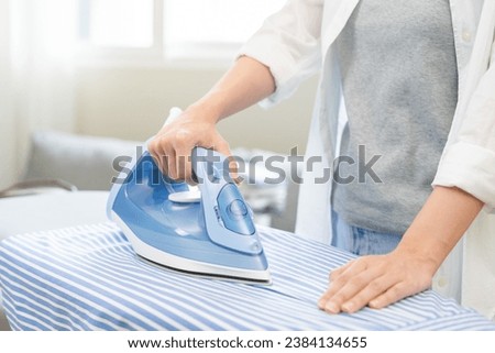 Close up young woman hand use electric steam, hot iron press pile stripe shirt clothes on ironing board, housework after hygiene laundry at home. Housekeeping lifestyle, household of chores concept. 商業照片 © 