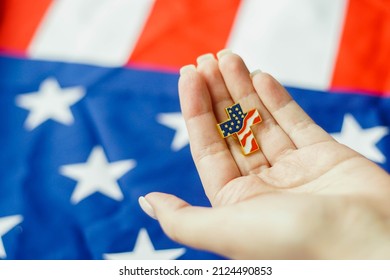 Close up of young woman holding Christian cross pin in her hand. American flag on the background. Patriotism and religious rights concept. - Powered by Shutterstock