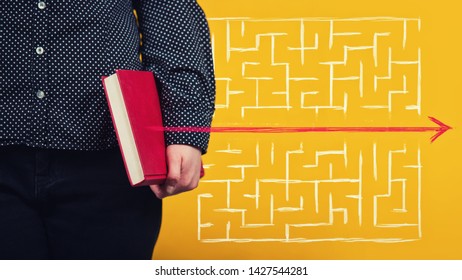 Close Up Of Young Woman Holding A Red Book As An Arrow Go Out And Pierce The Maze Walls. Education Concept As Knowledge Find Solutions To Escape From Labyrinth. Breaking The Rules, Different Thinking.