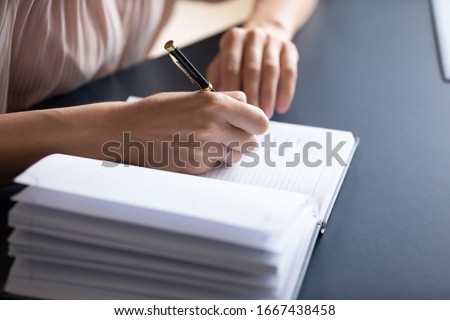 Close up young woman making notes in notebook, sitting at table. Female student listening to educational lecture, writing thesis in copybook. Successful businesswoman planning working day meetings.