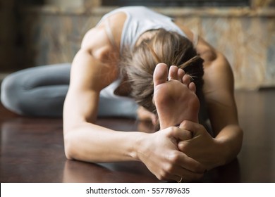 Close up of young woman practicing yoga, sitting in Head to Knee Forward Bend exercise, Janu Sirsasana pose, working out, wearing sportswear, grey pants, bra, indoor, home interior background