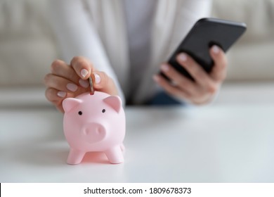Close up young woman putting coin in little piggy bank while managing investments, family budget or medical insurance payments in mobile banking application, personal savings expenditures concept.