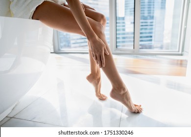 Close up young woman sitting on bath in modern bathroom with windows, touching legs, enjoying perfect smooth silky skin after epilation or home depilation procedure, applying moisturizing body cream