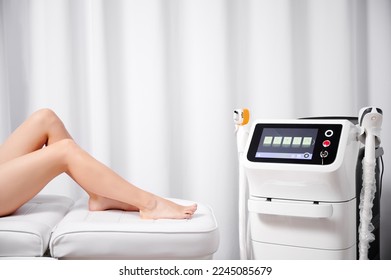 Close up of young woman sitting on daybed near diode laser hair removal machine in cosmetology clinic. Beautiful female legs and diode laser epilation machine in beauty salon.