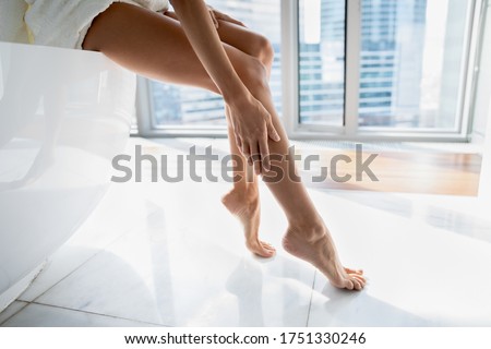 Close up young woman sitting on bath in modern bathroom with windows, touching legs, enjoying perfect smooth silky skin after epilation or home depilation procedure, applying moisturizing body cream