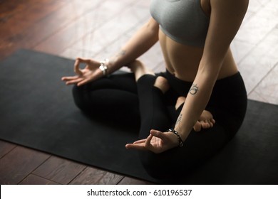 Close up of young woman with tattoo and nice wrist bracelets practicing yoga, sitting in Padmasana exercise, Lotus pose with mudra, working out on black mat, wearing sportswear, studio background 