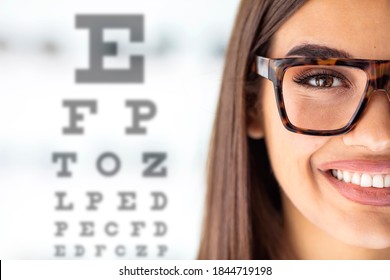 Close Up Of Young Woman Wearing Eyeglasses With Eyechart In The Background. Eyewear. Female Children's Doctor Near Eye Chart In Hospital. Young Woman On Eyesight Test Chart Closeup