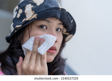 Close up of young woman wearing hat sitting outdoor. Beautiful Asian girl cleaning sweat on her face and using facial tissue on her face. Females are experiencing skin problems. - Shutterstock ID 2140157233