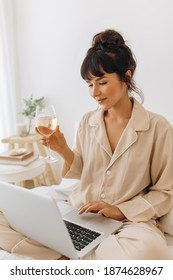 Close Up Of Young Woman Working On Laptop Computer Holding A Glass Of Wine. Portrait Of Woman In Night Suit Sitting On Bed At Home Using Laptop.