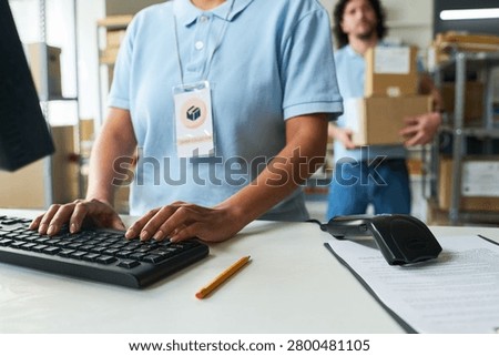 Close up of young worker processing pickup orders at distribution counter while coworker carrying parcels in background. Useful for logistics, delivery service, retail fulfillment