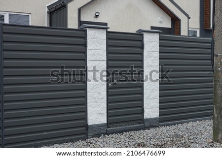 closed black metal door on a fence wall made of iron and white concrete pillars on a rural street