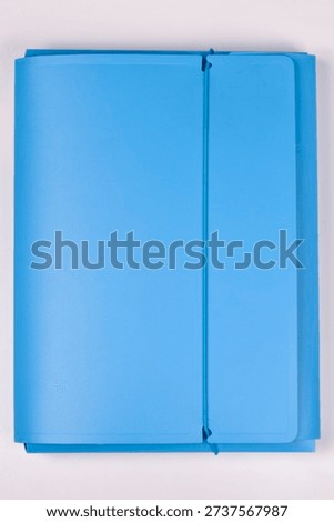 A closed blue plastic document folder secured with a vertical elastic band lies flat on a soft pink surface for office and school organization