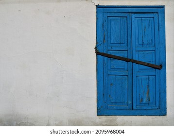 Closed Blue Wooden Shutters On The Wall Of An Abandoned House, Old Building With Cracked Stucco On The Facade And Textured Shutters With A Lock On The Awning