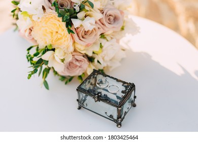 Closed Box With Metal Forging And Gold Rings For The Bride And Groom On A White Table With A Bouquet Of Wedding Flowers.