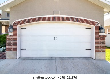 Closed Carriage Style White Garage Door Of A House With Bricks. There Are Two Wall Lamps On Either Side Of The Door And A Concrete Driveway To Garage.