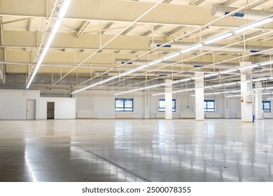 Closed down empty factory interior manufacturing hall with tall ceiling, concrete beams. Wide angle shot, ceiling mounted neon lights, no people.