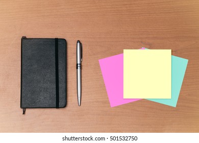 Closed Executive Notebook With Pen Metallic Finish On Table Textured Wooden Desk Three Post Its Color, Pink, Yellow And Blue, To Fill Gaps