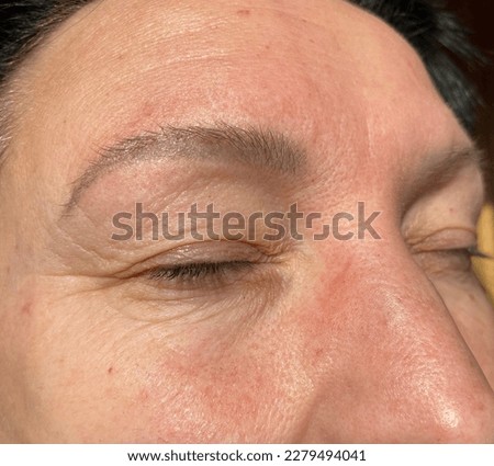 Closed eyes of a woman with mimic wrinkles in their natural form, without makeup. Eyebrows with a tattoo, sparse, uncombed hairs. Pores are visible on the face. Forehead with shallow wrinkles.