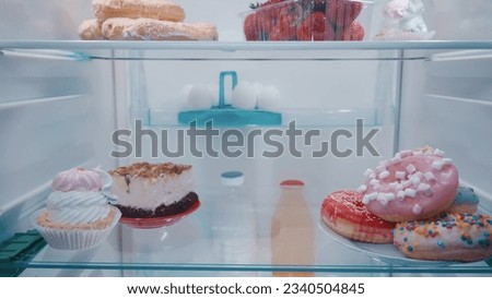 Closed fridge with unhealthy food, inside view. Pastries, eclairs, cake, donuts, strawberries, eggs, milk on the shelves in the refrigerator.
