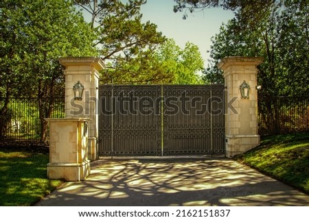 Closed impressive wrought iron luxury  security gate with key pad for private residential estate in leafy neighborhood with green trees and shadows on drive