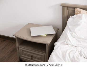 Closed laptop computer on a bedside table next to a bed with crumpled white sheets, representing technology in a relaxed home setting