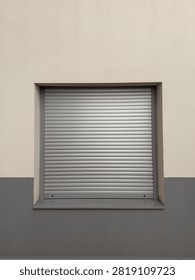A closed light gray roller shutter covering a square window on a two-tone stucco house facade. Exterior aluminum roller blinds for sun protection and privacy.