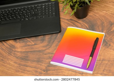 closed notebook with a bright colorful gradient on the cover and a black pen lie on a wooden table near the keyboard of an open laptop and a green indoor flower in a pot