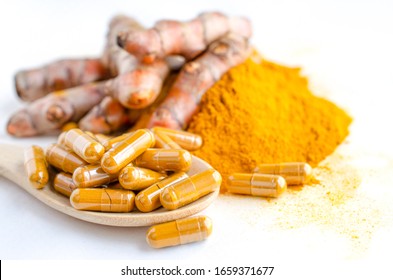Closed up organic turmeric or curcumin powder in capsule on wooden spoon over blur background of fresh turmeric plant and powder pile on white background,for Thai herb or traditional medicine concept