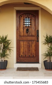 Closed Ornate Wood Door Of An Upscale Home, Accented With An Iron Barred Window, Iron Door Knocker, And Iron Bolts. The Door Is Set Into A Beige Stucco House, Framed With A Wide Arch And Planters.