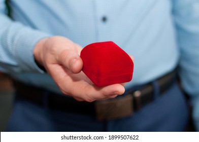 A Closed Red Velvet Ring Box In The Hands Of A Man In A Blue Shirt